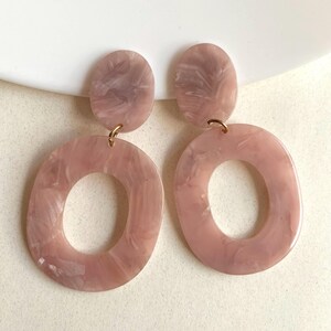 May include: A pair of drop earrings featuring a small oval top piece connected to a larger, open oval bottom piece. Both components are made of a light pink, marbled acrylic material with a glossy finish, joined by gold-toned metal jump rings.