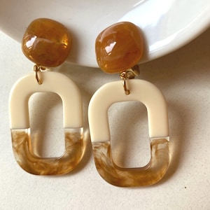 May include: A pair of drop earrings featuring a smooth, amber-coloured resin stud connected by a gold-tone metal jump ring to a rectangular pendant. The pendant is composed of an opaque cream-coloured top and a translucent, marbled amber base.