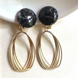 May include: A pair of clip-on earrings featuring a circular black and white marbled stud top connected to a gold-toned metal pendant. The pendant consists of multiple thin, overlapping oval-shaped wire loops that create a layered, open-frame design.