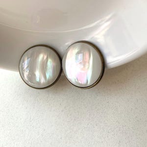 May include: Two round, iridescent white earrings with a bronze-colored metal border. The earrings are set against a white background, showcasing their pearlescent sheen. These earrings are a classic accessory.