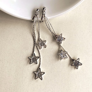May include: Silver-toned dangle earrings featuring a star design. Each earring has a small ball stud and multiple strands of delicate chain, each ending with a star-shaped charm with a clear center. The earrings are displayed on a white surface.