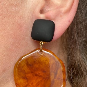 May include: A pair of statement earrings featuring a black square-shaped top and a large, round, amber-coloured bottom. The earrings have a gold-coloured connector between the top and bottom pieces.