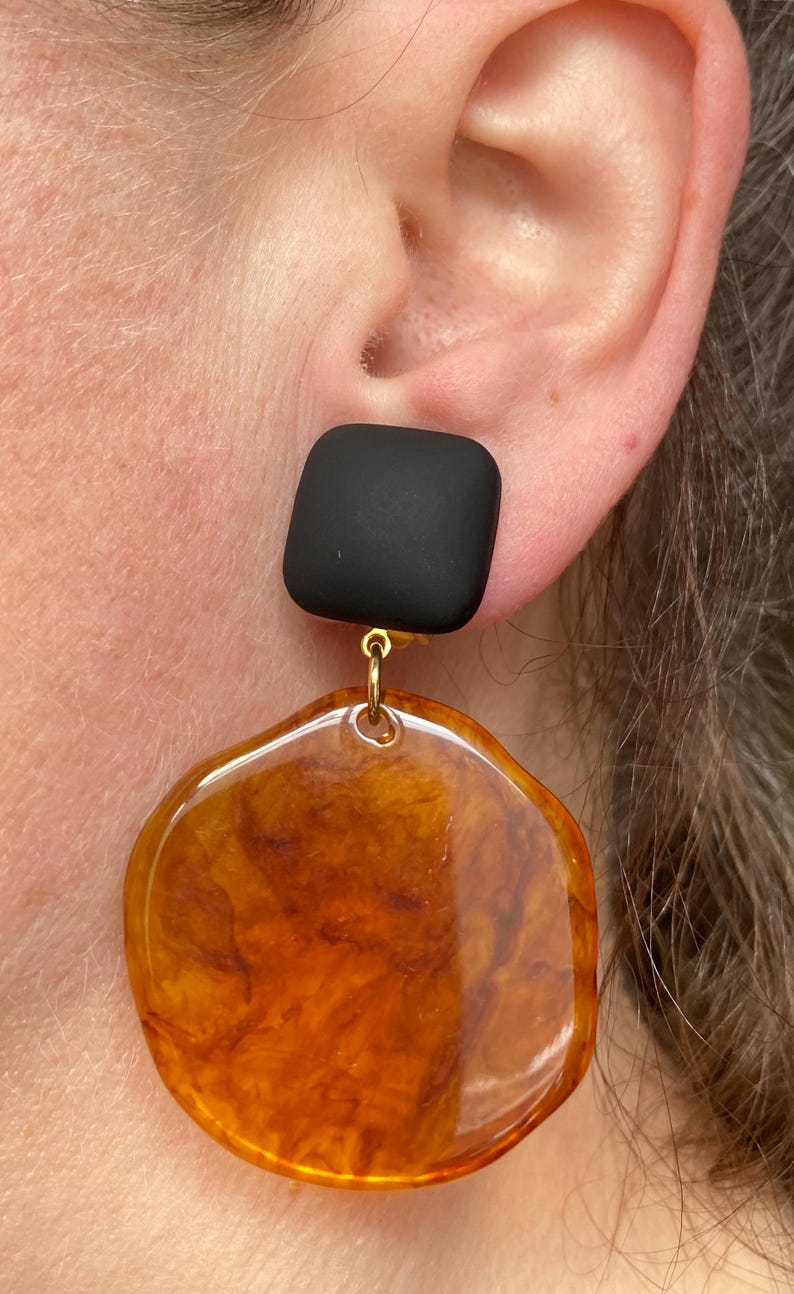 May include: A pair of statement earrings featuring a black square-shaped top and a large, round, amber-coloured bottom. The earrings have a gold-coloured connector between the top and bottom pieces.