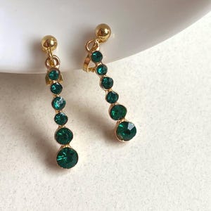 May include: A pair of gold-toned earrings with a vertical cascade of emerald green gemstones. Each earring features a small gold ball at the top and a larger gemstone at the bottom. The gemstones are round and faceted, reflecting light.