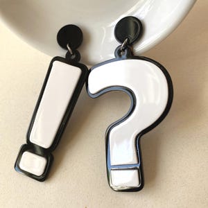 May include: A pair of quirky earrings, one shaped as a white exclamation mark and the other as a white question mark, both with black outlines. They have black circular studs and are set against a plain background.