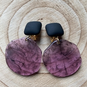 May include: A pair of black and purple clip-on earrings. The earrings feature a black square-shaped top and a large, round, translucent purple bottom with a marbled pattern.