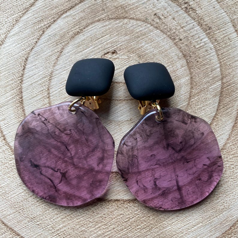May include: A pair of black and purple clip-on earrings. The earrings feature a black square-shaped top and a large, round, translucent purple bottom with a marbled pattern.