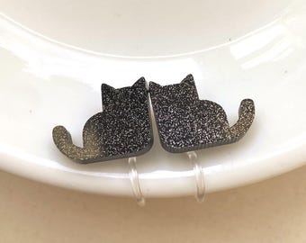 May include: A pair of black cat-shaped earrings with a glittery finish. The earrings are attached to clear, plastic earring clips. The cat silhouettes are sitting with their tails curved upwards. The earrings are on a white surface.