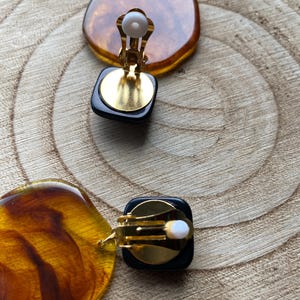 May include: A pair of vintage clip-on earrings with a black and amber resin design. The earrings have a gold-tone metal clip closure.