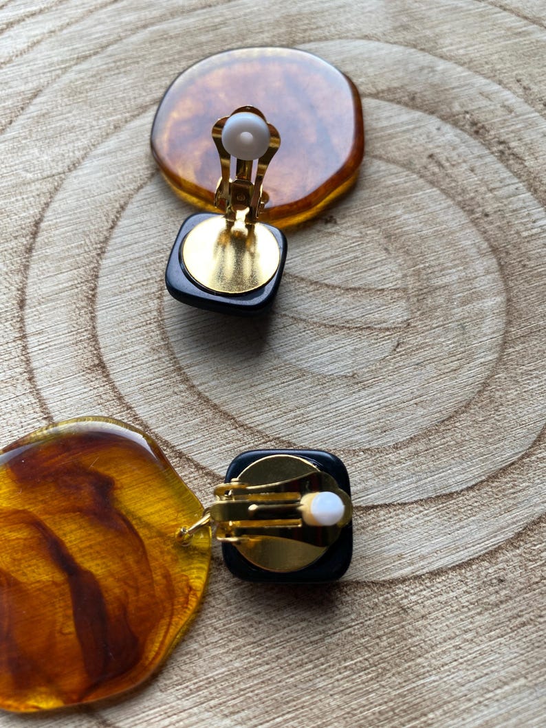 May include: A pair of vintage clip-on earrings with a black and amber resin design. The earrings have a gold-tone metal clip closure.
