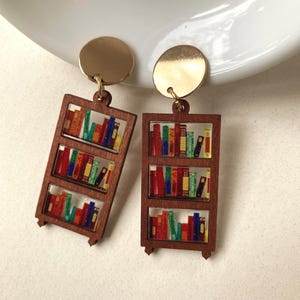 May include: A pair of earrings with a bookshelf design. Each earring has a gold-coloured circular top and a wooden frame with three shelves filled with colourful, miniature books. The earrings are designed to dangle.