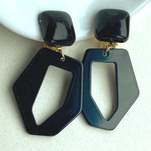 May include: A pair of dark blue geometric earrings. Each earring features a square top and a large, open, diamond-shaped pendant. The earrings are connected with gold-colored hardware. The earrings are displayed on a white surface.