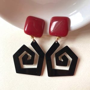 May include: A pair of statement earrings. They have square, burgundy tops and black geometric pendants with a spiral design. The earrings have gold-coloured clasps. Set against a white background.