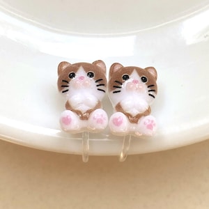 May include: A pair of cat-shaped stud earrings featuring brown and white resin figures with black eyes, pink noses, and pink paw prints on the feet. The earrings are attached to clear plastic clip-on components for secure wear without piercings.