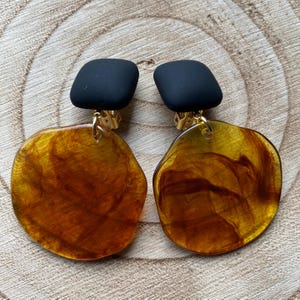 May include: A pair of clip-on earrings with black square tops and large, round, amber-coloured bottoms with a marbled pattern.