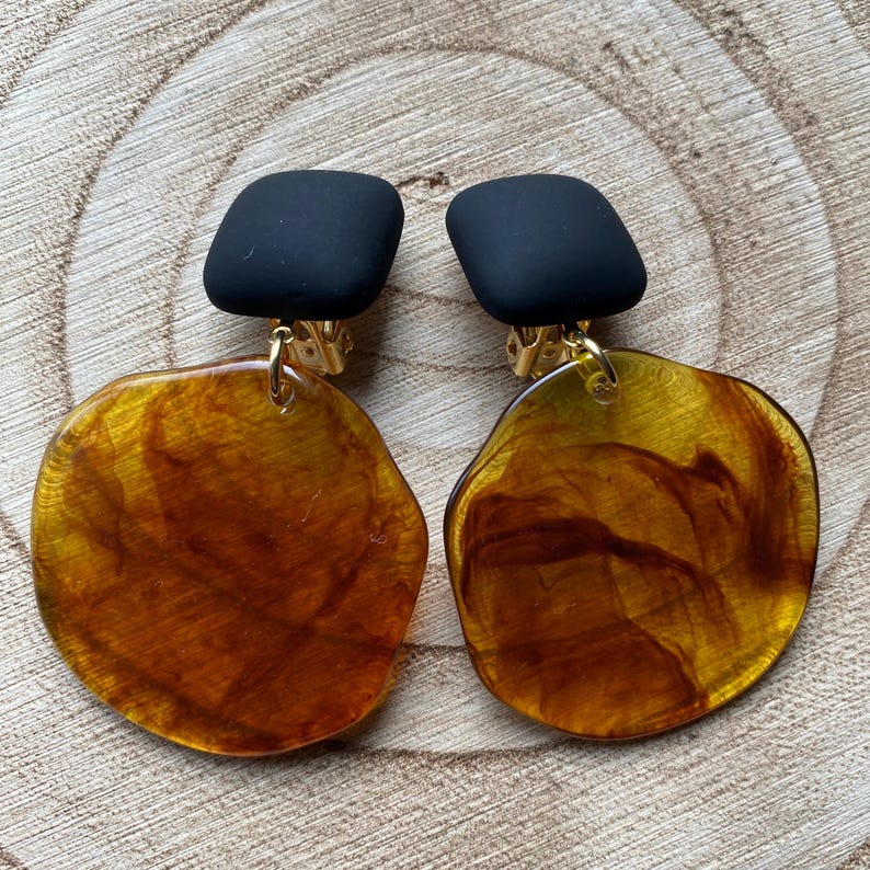 May include: A pair of clip-on earrings with black square tops and large, round, amber-coloured bottoms with a marbled pattern.