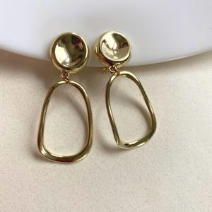 May include: A pair of gold-coloured earrings. Each earring has a circular stud connected to a teardrop-shaped open frame. The earrings are displayed on a white surface, highlighting their polished, reflective finish.