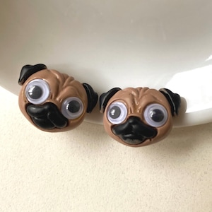 May include: Two brown pug-shaped embellishments with black ears and snouts, and large, googly eyes. The pug faces are slightly different, with one angled to the left. The background is a white, slightly curved surface.