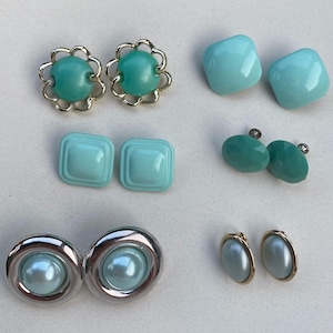 May include: A collection of six pairs of turquoise and light blue earrings. The set includes floral-shaped metal settings, smooth square studs, faceted round studs, silver-toned circular frames with light blue centres, and oval-shaped studs with gold-toned rims.