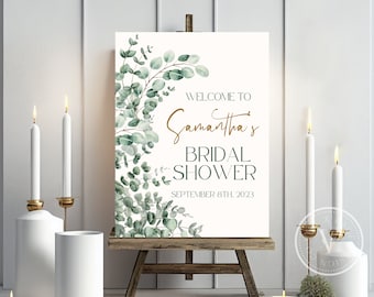 May include: A white sign with green eucalyptus leaves and gold lettering. The sign says "Welcome to Samantha's Bridal Shower September 8th, 2023".
