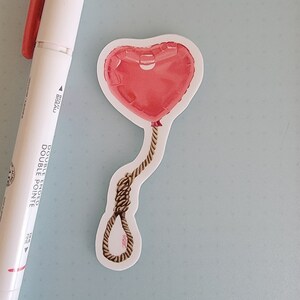 Puede incluir: Un globo en forma de corazón de color rojo está atado a una cuerda marrón, formando un nudo corredizo. El globo y la cuerda están sobre un fondo blanco.