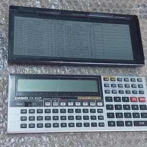 May include: A black and silver Casio FX-850P scientific calculator with a small LCD screen displaying an error message table. The calculator has a variety of buttons, including numbers, operations, and functions. The calculator is a vintage model and is in good condition.