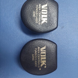 May include: Two black round cases with gold lettering. The text on the cases reads "VOLK AREA CENTRALIS FUNDUS/LASER LENS" and "VOLK AREA CENTRALIS FUNDUS/LASER LENS ®".