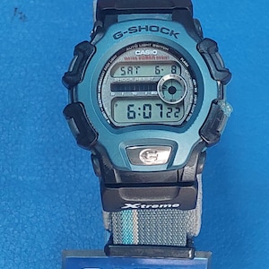 May include: A blue Casio G-Shock digital watch with a black band and a grey strap. The watch face displays the time, date, and day of the week. The watch is water resistant and shock resistant. The watch has a black and grey Xtreme logo on the strap.