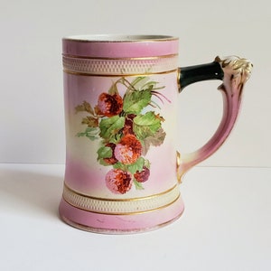 Op de afbeelding: Een vintage keramische bierpul in roze en crème. Het heeft een bloemenontwerp met rode bessen en groene bladeren. De handgreep is donkergroen, met een leeuwenkopdetail. Gouden randen accentueren het ontwerp.