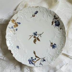 May include: White porcelain serving plate with a scalloped edge and gold trim. The plate features embossed floral designs and is decorated with blue and gold floral motifs. A decorative piece for serving food or displaying.
