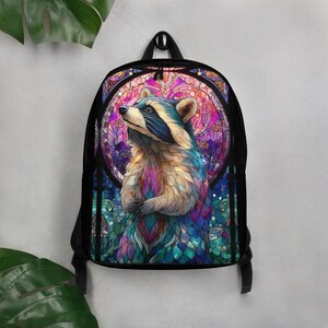 Könnte beinhalten: Ein schwarzer Rucksack mit einer zentralen Grafik eines Waschbären im bunten Buntglas-Stil. Der Waschbär ist im Profil vor einem kreisförmigen, mehrfarbigen geometrischen Hintergrund in Rosa-, Lila- und Blautönen dargestellt.