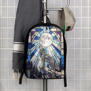 May include: A black backpack featuring a graphic print of a wolf howling at a full moon, designed with a stained glass aesthetic in shades of blue, purple, and white. The backpack has black straps and a black frame, showcasing a detailed scene.