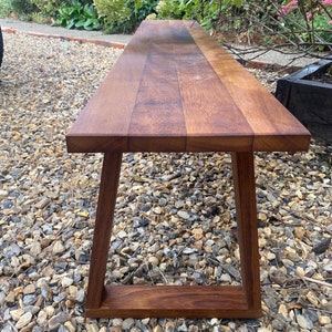 May include: A long wooden bench with a natural brown finish and a simple, modern design. The bench has a rectangular top and a frame with angled legs.