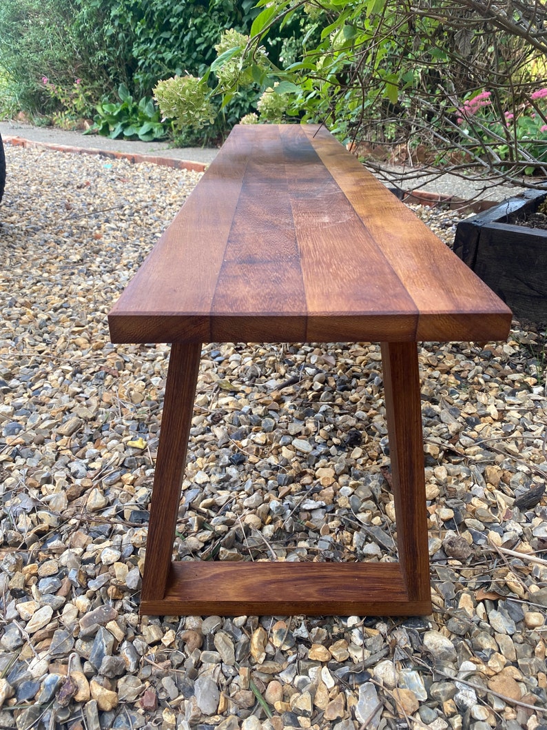 May include: A long wooden bench with a natural brown finish and a simple, modern design. The bench has a rectangular top and a frame with angled legs.