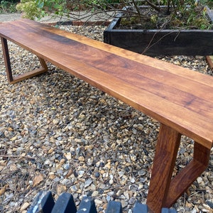 May include: A long, wooden bench with a natural finish. The bench has a simple, modern design with a rectangular top and angled legs. The bench is made of solid wood and has a warm, natural color.