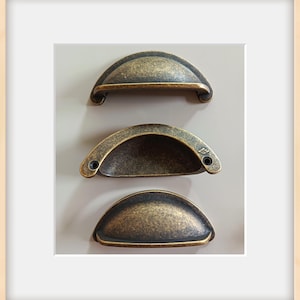 May include: Three antique-style cabinet pulls with a bronze finish. Each pull has a half-moon shape with a textured surface and two screw holes. The pulls are arranged vertically against a neutral background, showcasing their design.