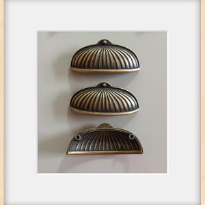 May include: Three antique-style drawer pulls with a bronze finish. Each pull has a half-circle shape with a ribbed design. The pulls are mounted on a light gray surface, suggesting a cabinet or furniture application.