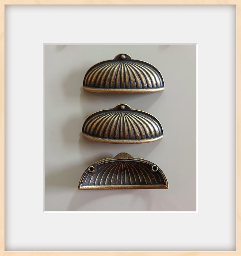 May include: Three antique-style drawer pulls with a bronze finish. Each pull has a half-circle shape with a ribbed design. The pulls are mounted on a light gray surface, suggesting a cabinet or furniture application.