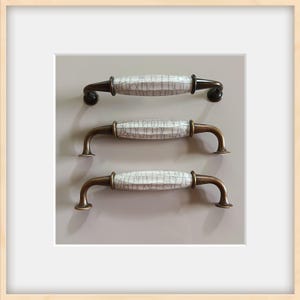 May include: Three cabinet handles with a white, crackled ceramic center and bronze-colored metal hardware. The handles are arranged vertically against a neutral gray background, showcasing their design and potential use in home decor.