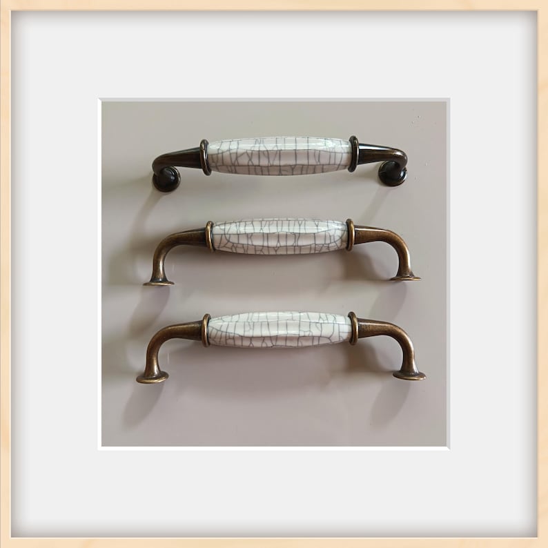 May include: Three cabinet handles with a white, crackled ceramic center and bronze-colored metal hardware. The handles are arranged vertically against a neutral gray background, showcasing their design and potential use in home decor.