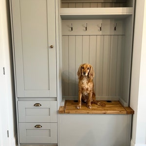 May include: A light grey mudroom storage unit featuring a tall cabinet with a single door, two lower drawers with brass-toned cup pulls, and an open bench area with a natural wood seat, four metal coat hooks, and a matching wood top shelf.