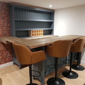 May include: A bespoke home bar with a grey-blue base and a wooden countertop. It features three brown leather swivel bar stools with black bases. The bar has a built-in shelving unit and a wine rack.