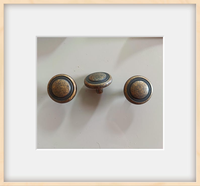 May include: Three antique bronze cabinet knobs with a circular design. Each knob features a raised center and a dark ring around the edge. The knobs are set against a light background.