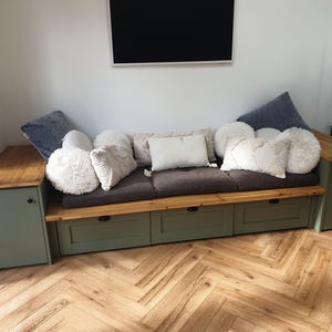 May include: A bespoke sofa with a dark brown seat cushion and various decorative pillows. The sofa features a green base with drawers and wooden armrests. The room has a wooden floor and a wall-mounted TV.