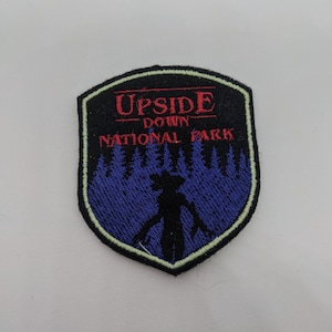Könnte beinhalten: Ein schwarzer gestickter Aufnäher mit weißem Rand. Der Aufnäher zeigt die Silhouette eines Monsters vor einem violetten Wald mit dem Text "Upside Down National Park" in Rot.
