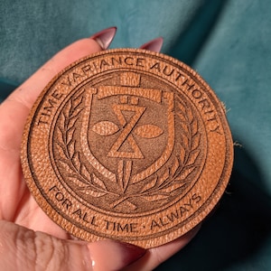 Könnte beinhalten: Ein braunes Lederabzeichen mit dem Text "Time Variance Authority" in einem Kreis um einen Schild mit einem Schwert und dem Buchstaben "Z" darin. Der Text "For All Time. Always" befindet sich unter dem Schild.