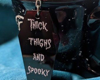 Puede incluir: Llavero negro en forma de ataúd con una cadena plateada. El llavero tiene texto blanco que dice "Thick Thighs and Spooky Vibes".