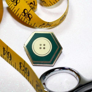 Könnte beinhalten: Eine goldene sechseckige Anstecknadel mit grünem Rand und einem weißen Knopf mit vier Löchern in der Mitte. Die Anstecknadel ist von einem gelben Maßband und einer silbernen Schere umgeben.