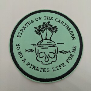 May include: A green and black embroidered patch with a skull and crossbones, palm trees, and the text "Pirates of the Caribbean Yo Ho A Pirates Life For Me".
