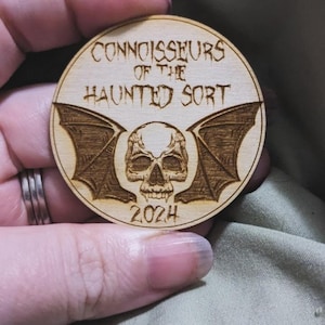 May include: A wooden round ornament with the text "Connoisseurs of the Haunted Sort" and a skull with bat wings. The year 2024 is also engraved on the ornament.